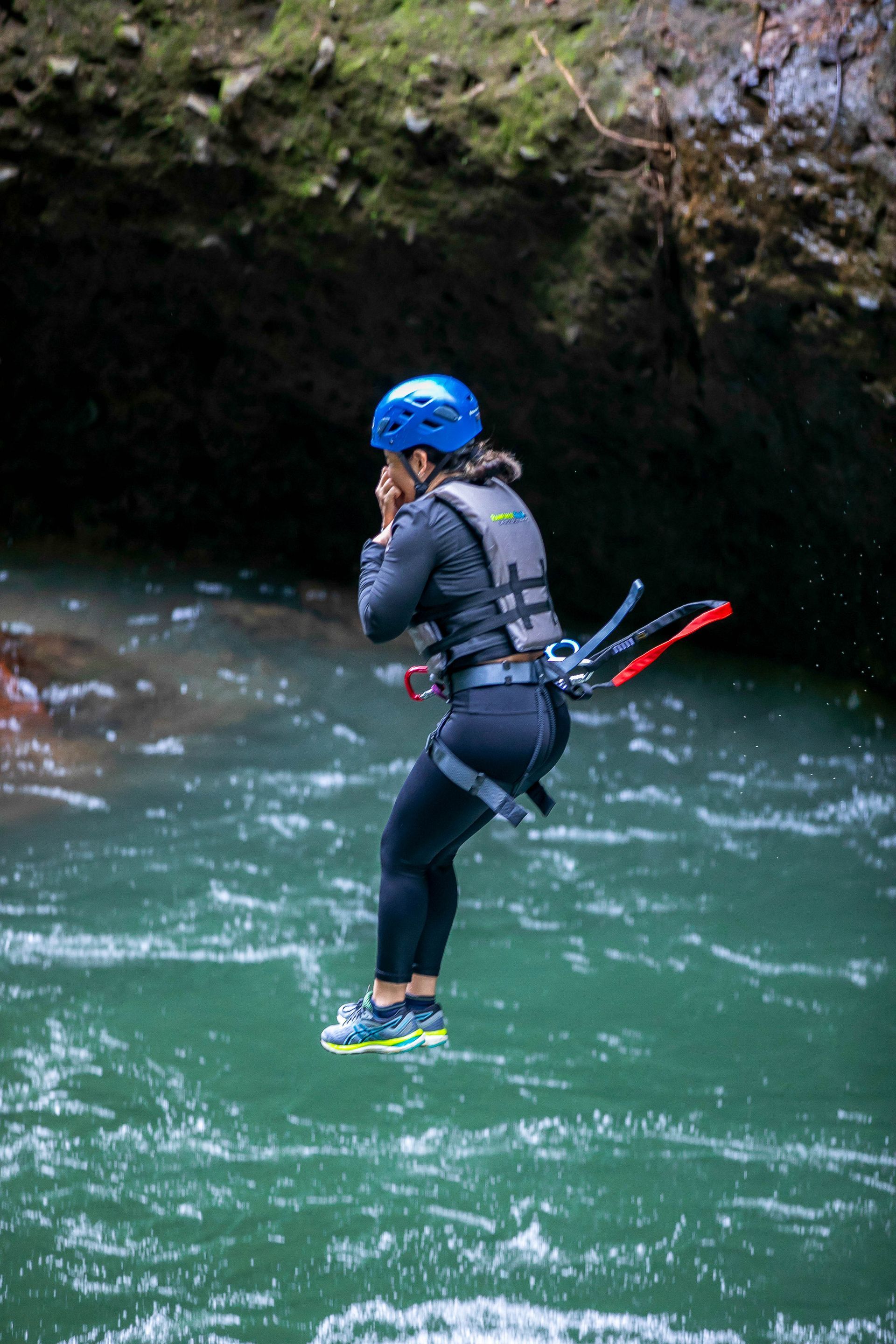Woman leaps into turquoise water, wearing helmet, life vest, and harness, under mossy cliff.