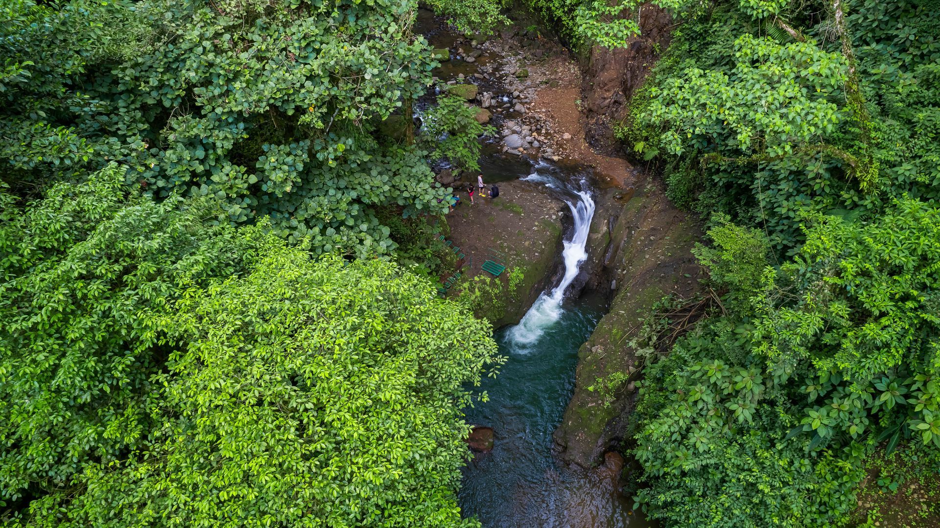 Aerial view: Waterfall cascading into a river, surrounded by lush green trees.