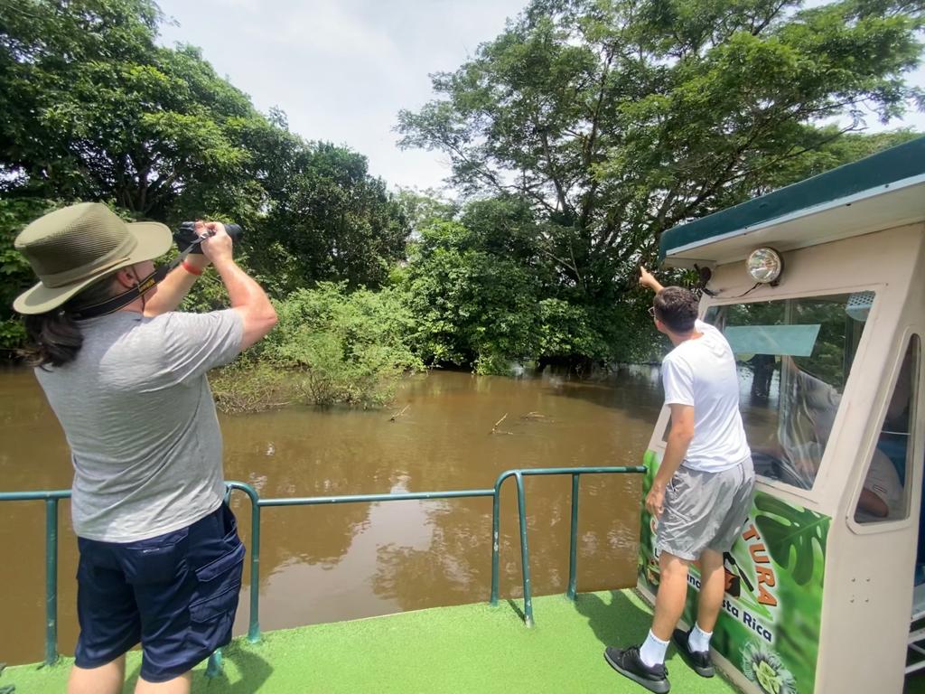 Two people on a boat taking photos of wildlife in a murky river, surrounded by green trees.