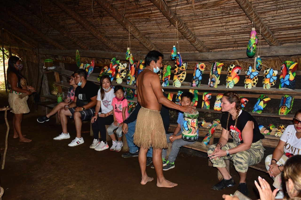 People in a hut watch a bare-chested man present colorful carvings.