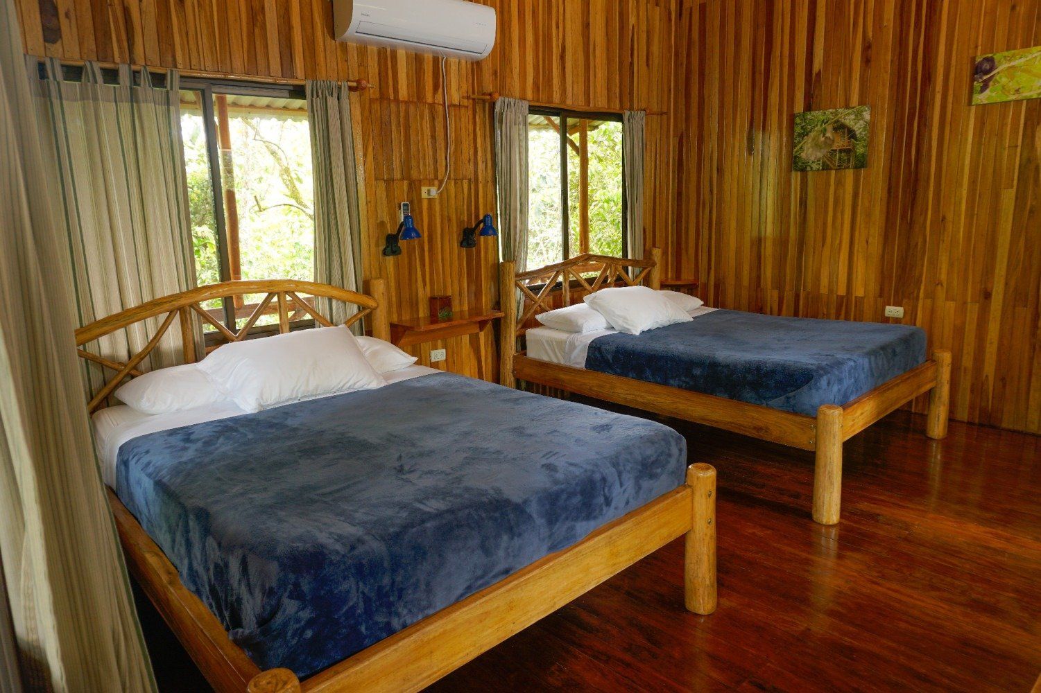 Two beds in a wood-paneled room with windows, blue blankets, and air conditioning.