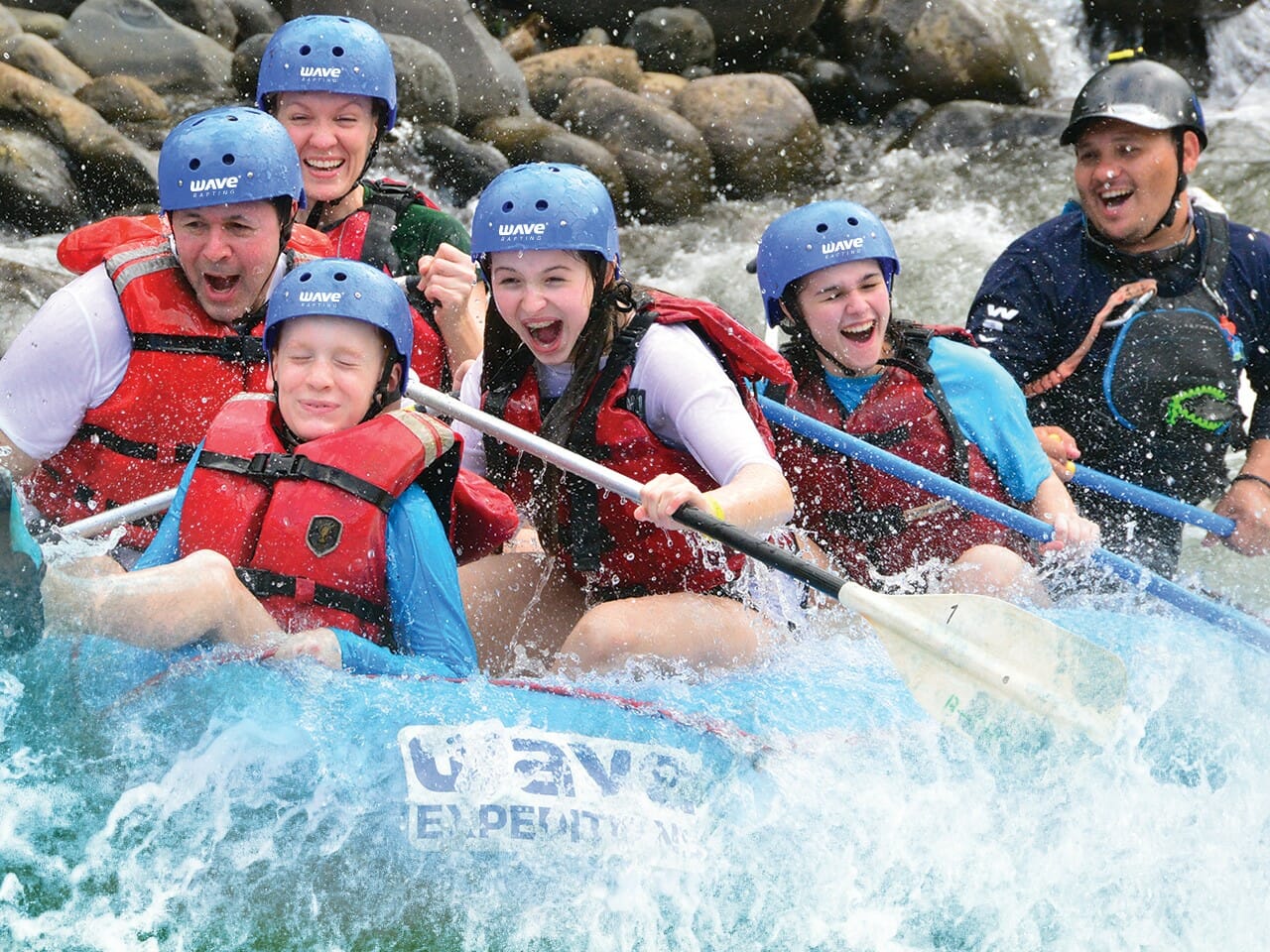 People whitewater rafting, laughing and paddling. Blue water, life vests, helmets.