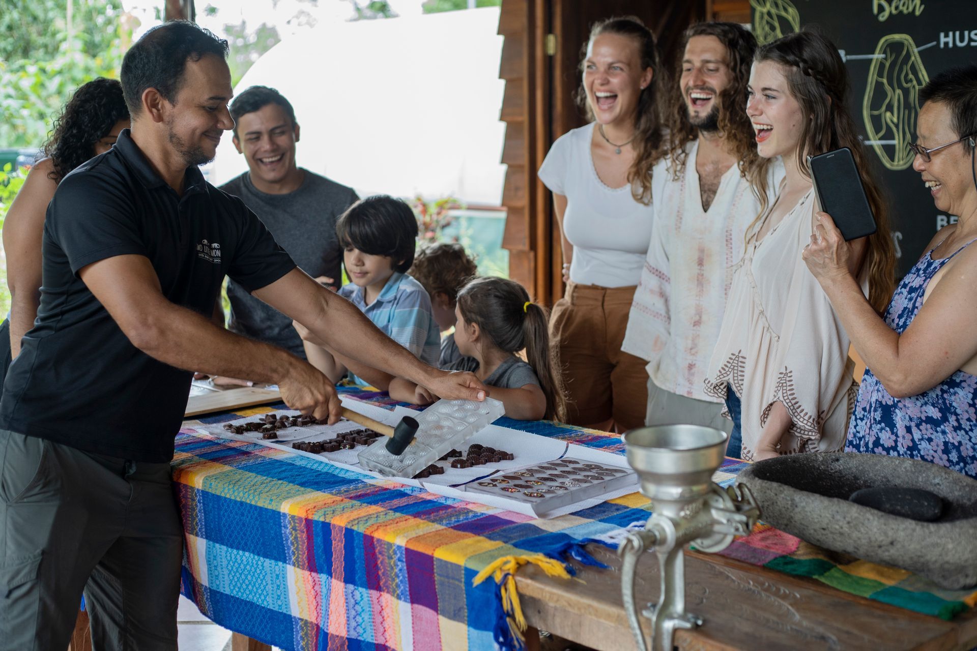 Group excited at a chocolate-making demonstration. Man points at table with chocolate, colorful tablecloth, chalkboard, and mortar.