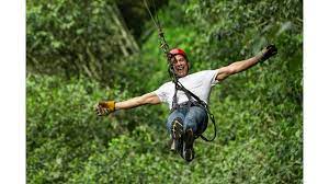 Man ziplining through a green forest, arms outstretched, smiling. Wearing helmet, harness, and gloves.