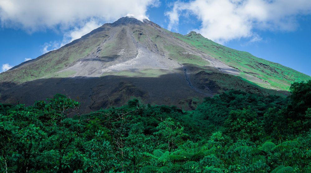 Green mountain with dark slopes rising above a lush, green forest under a blue sky.