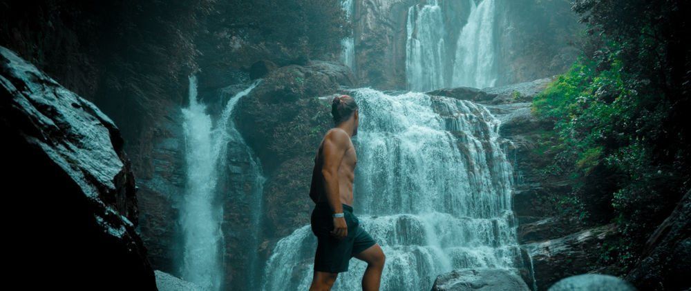 Man stands shirtless near a waterfall, looking at cascading water. Lush green foliage surrounds.