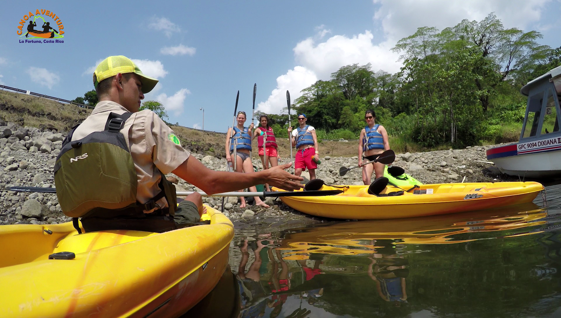 People kayaking near the shore; a guide in a scout uniform instructs others as they prepare to paddle.