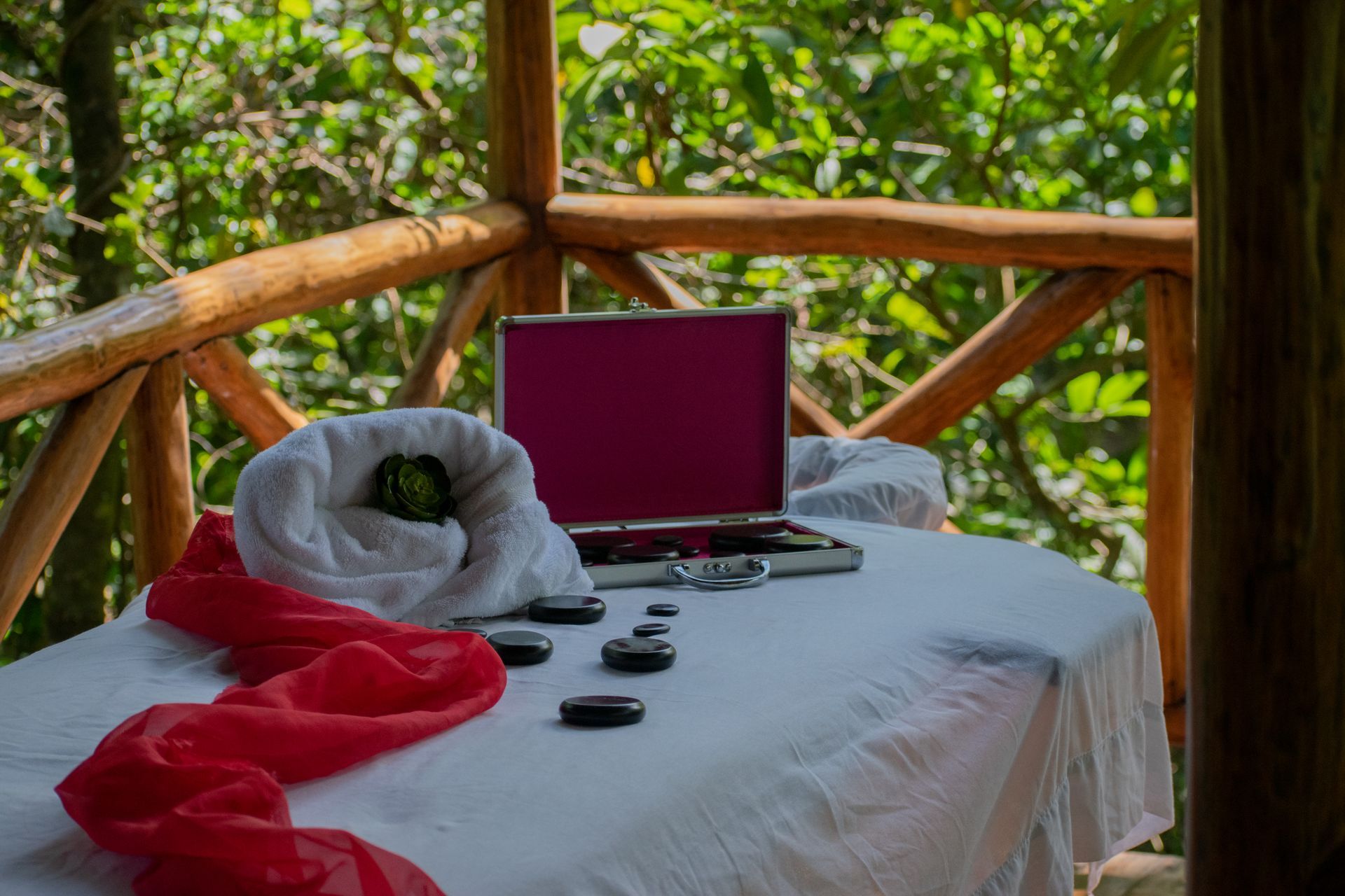Massage table on a wooden balcony overlooking green foliage, with red scarf, towels, and tools.