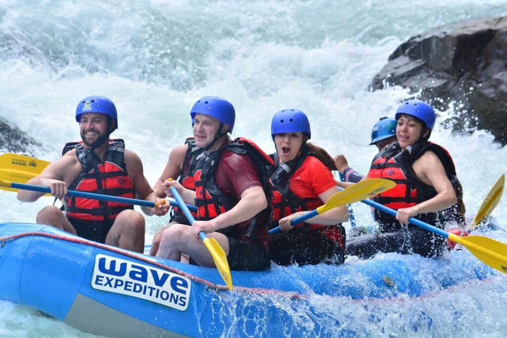 People white water rafting in a blue raft, wearing helmets and life vests, navigating rapids.