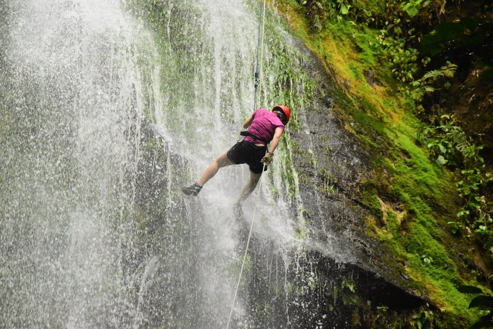 Person rappels down a waterfall, wearing a helmet and safety gear. Lush green vegetation and water spray surround them.