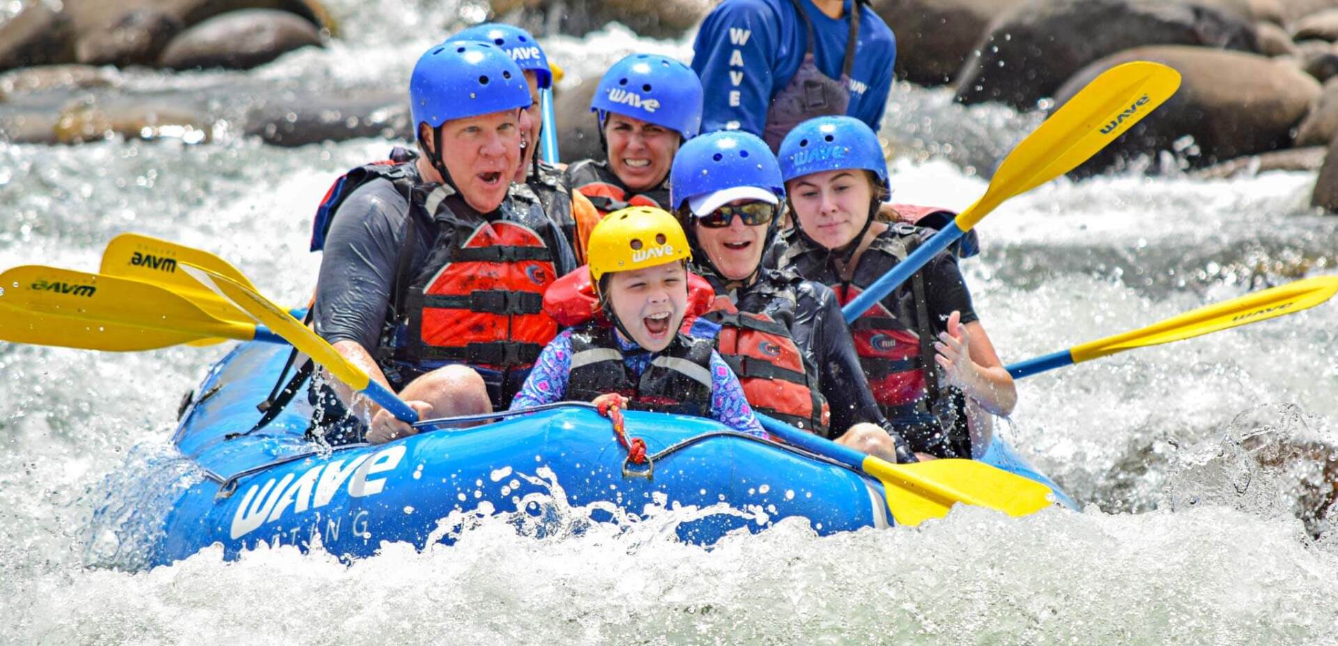 People whitewater rafting in a blue raft, wearing helmets and life vests. They are splashing in white water.
