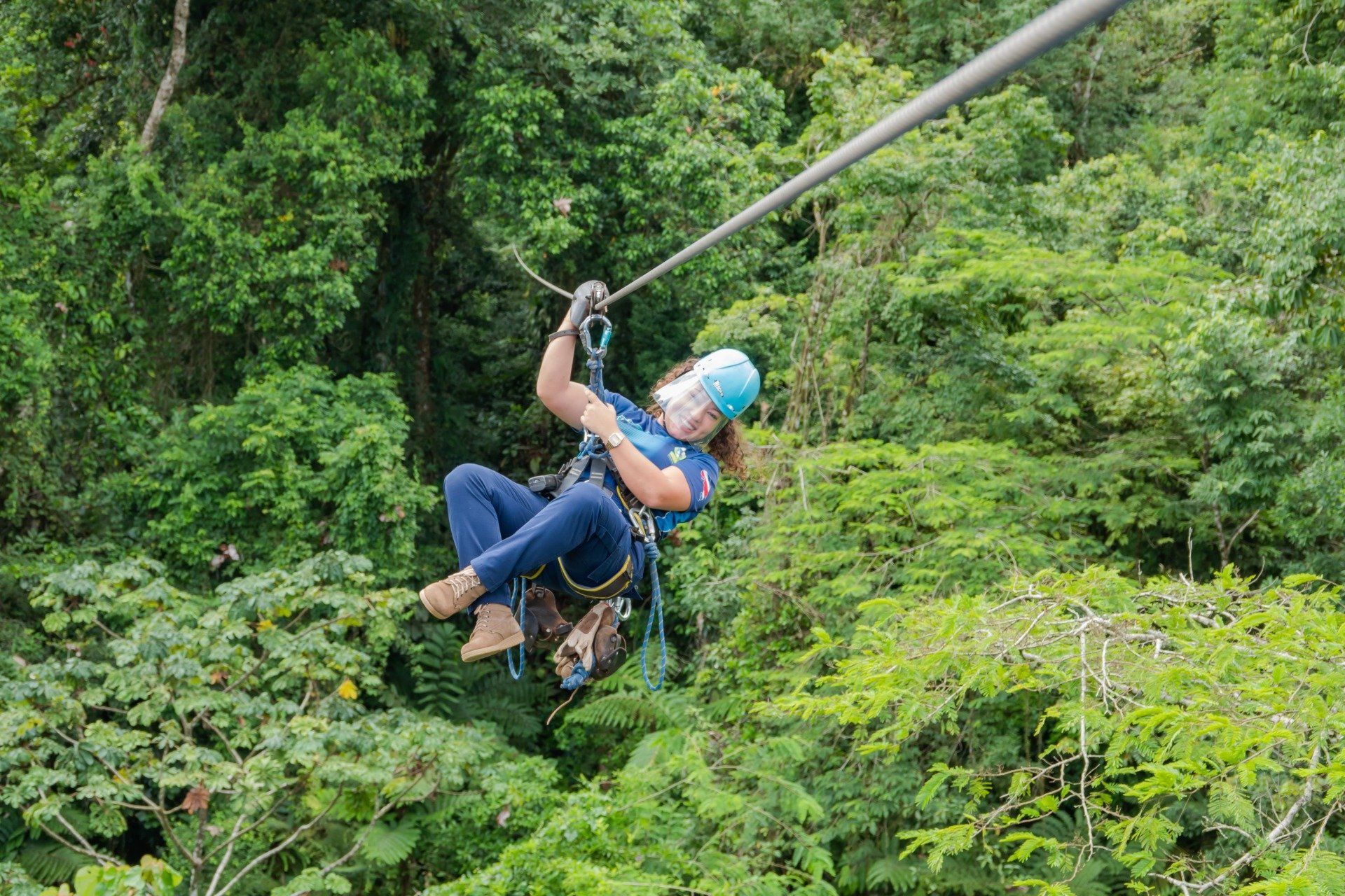 Person zip-lining through lush green rainforest, wearing a helmet and harness.