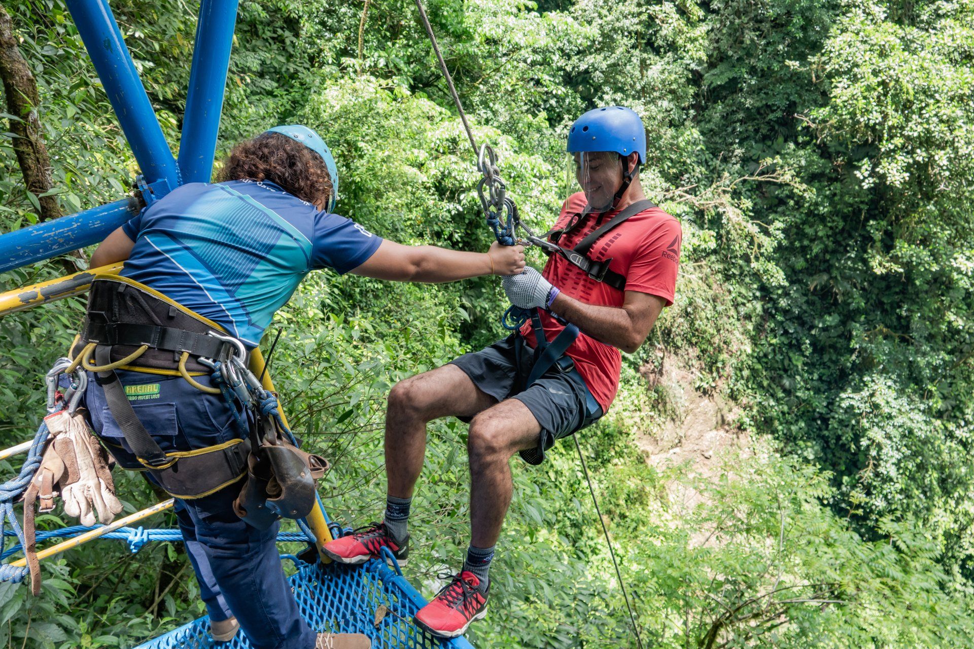 Man assisted on a zip line platform; green forest backdrop.