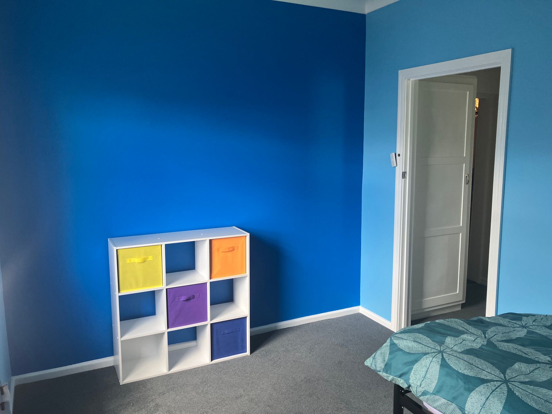 Empty blue bedroom with a white cubby shelf and an open door next to a bed with teal bedding.