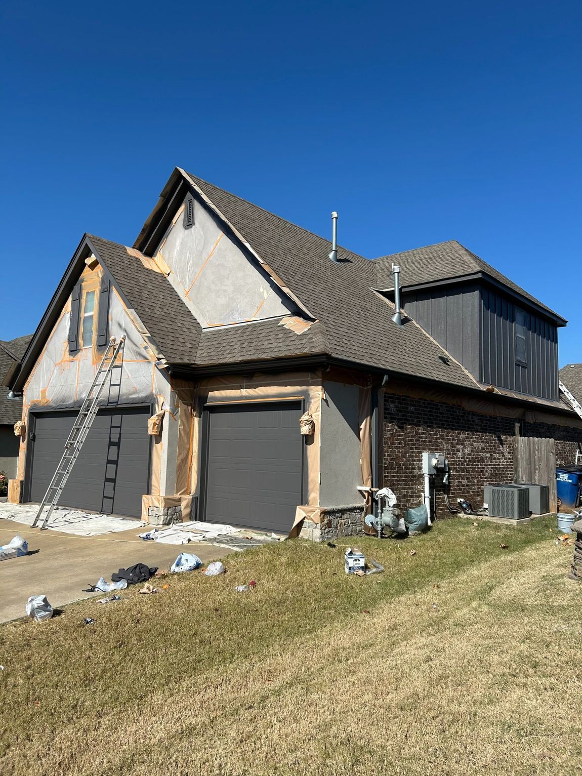 New house under construction with gray siding, stone accents, and a steep roof on a grassy slope under a blue sky