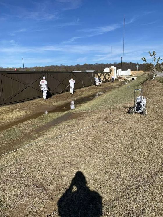People in white suits working along a muddy drainage ditch beside a tall wooden fence on a sunny day