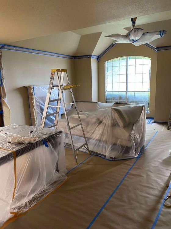 Living room under renovation, furniture covered in plastic, with ladder and taped-off walls.