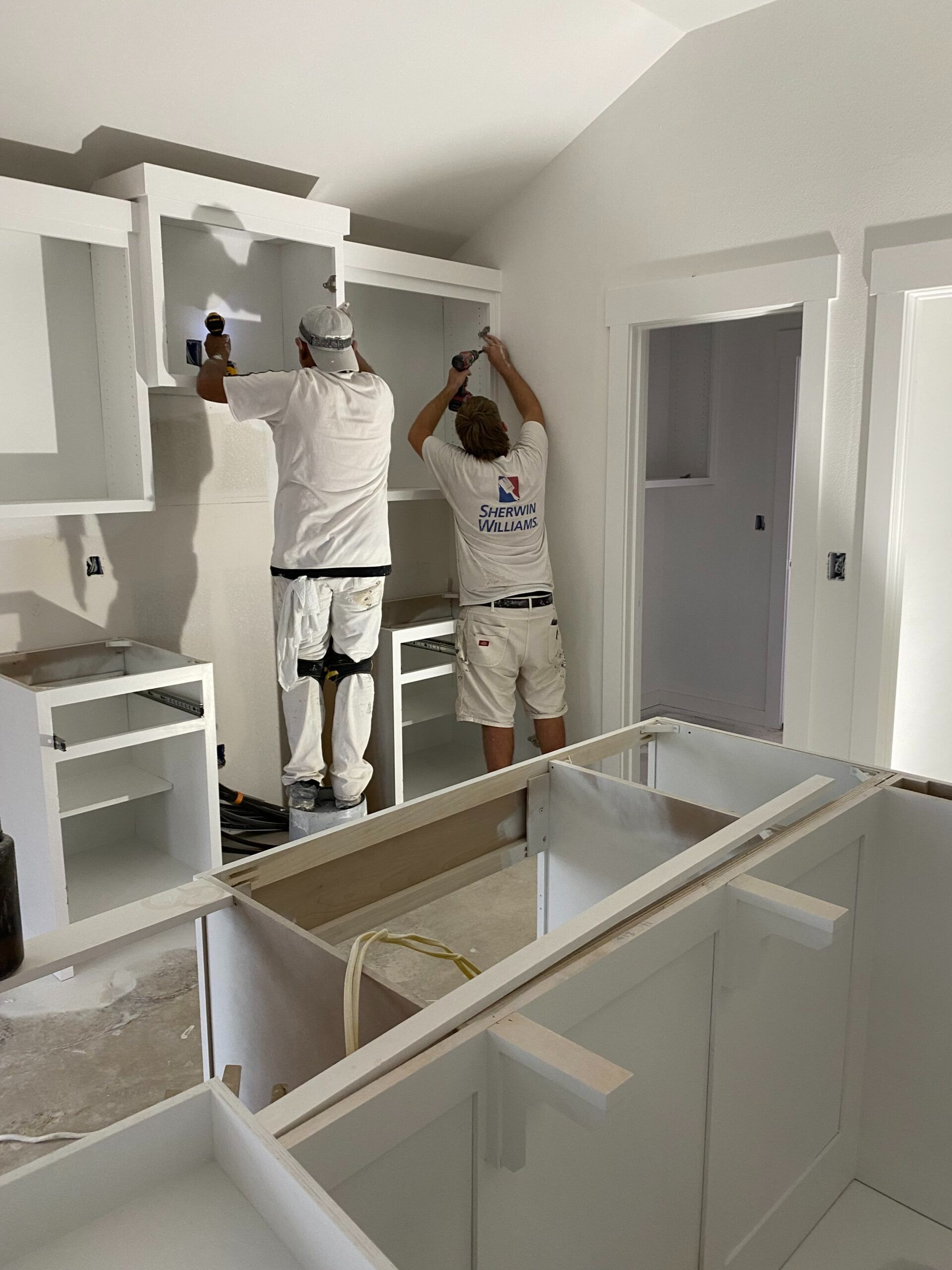 Two people painting white kitchen cabinets in an unfinished kitchen with exposed cabinet frames.