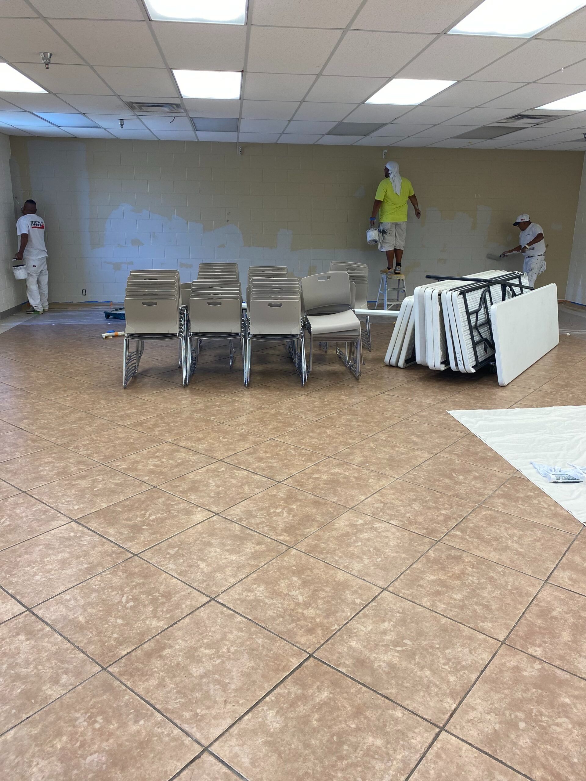 Empty room with stacked chairs, folded tables, and workers painting walls under a tiled ceiling