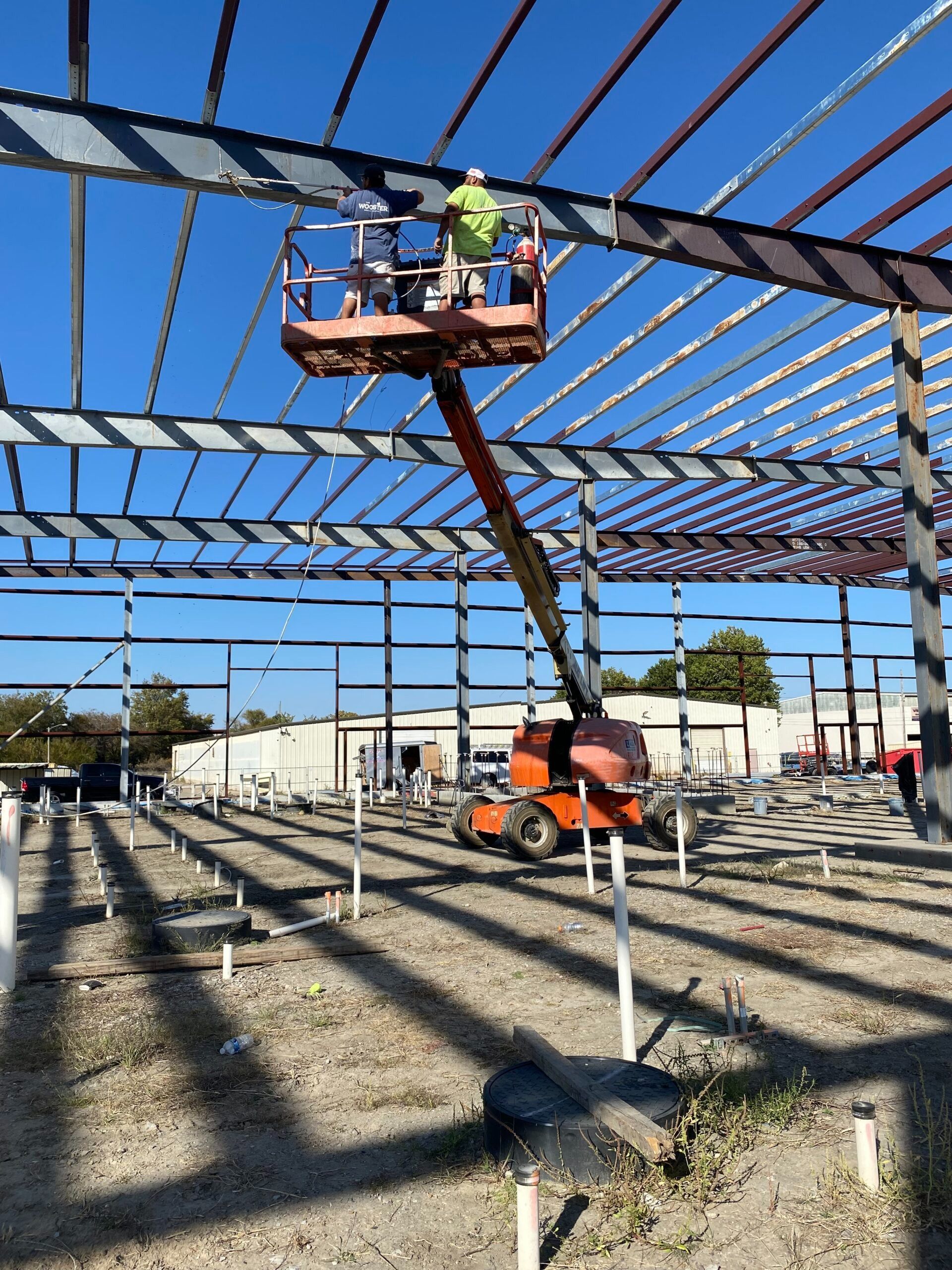 Workers on a lift installing roof framing on a steel building skeleton under a blue sky
