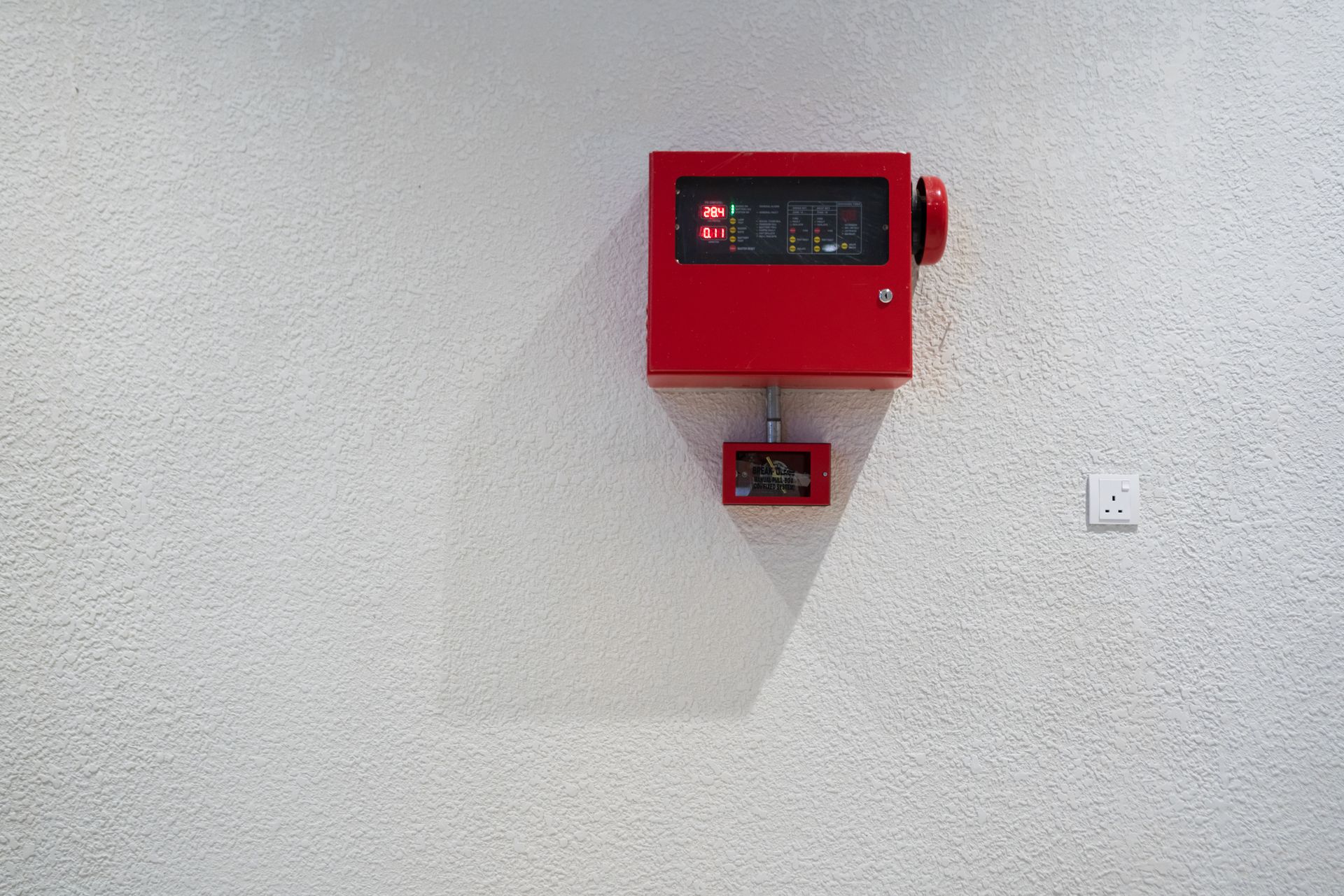 A man is fixing a fire alarm box with a screwdriver. Red fire alarm system on a textured white wall, with a small pull station below.