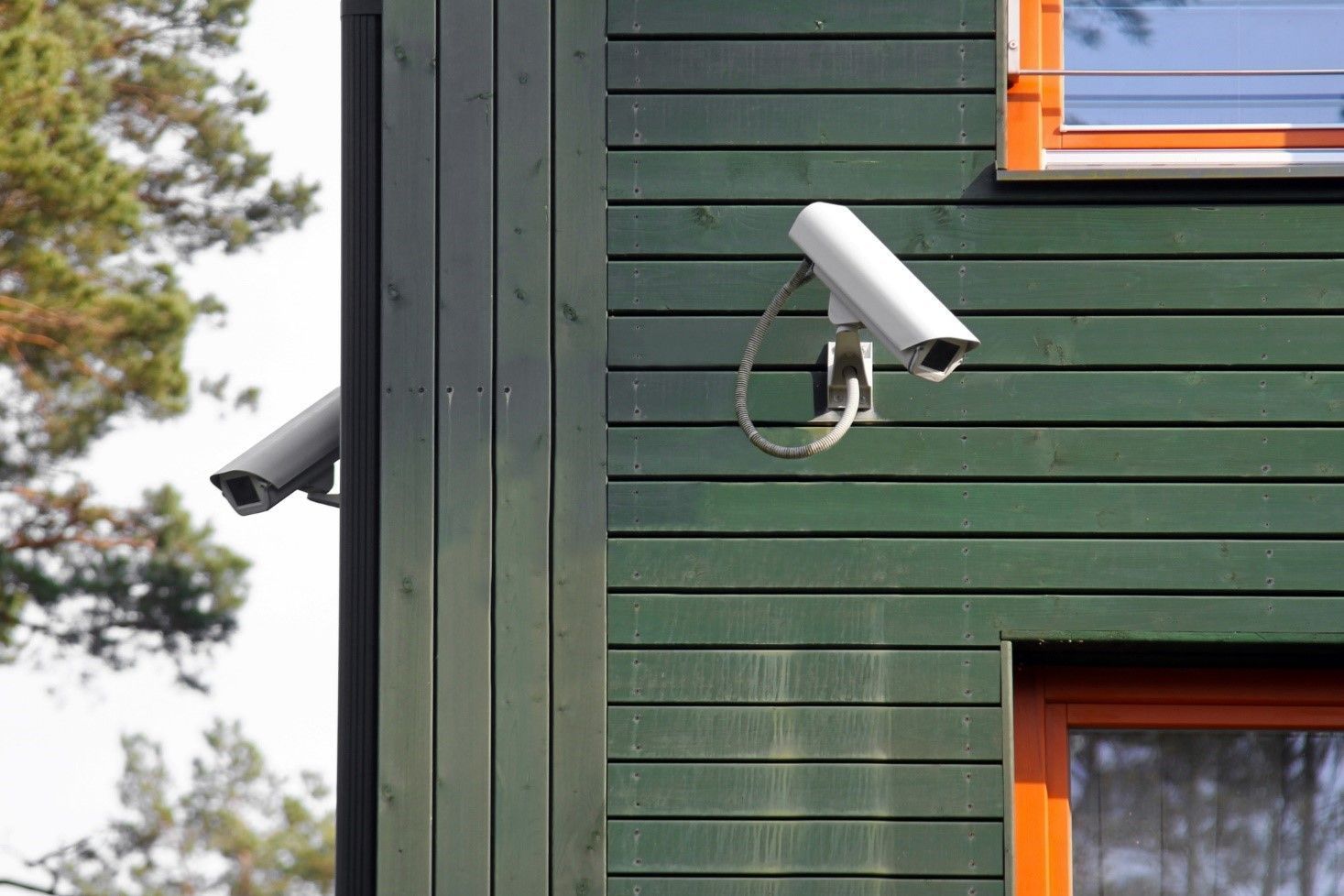 Two white security cameras mounted on a green wooden building corner, with a window visible.