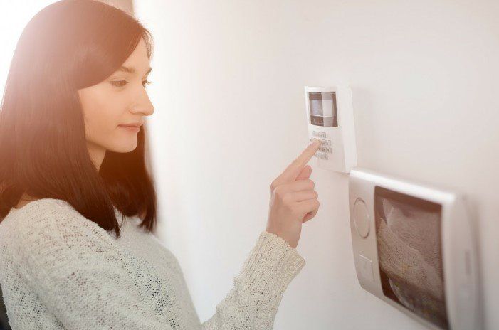 Woman in a sweater pressing a button on a white wall-mounted thermostat.  Another panel is below the thermostat.