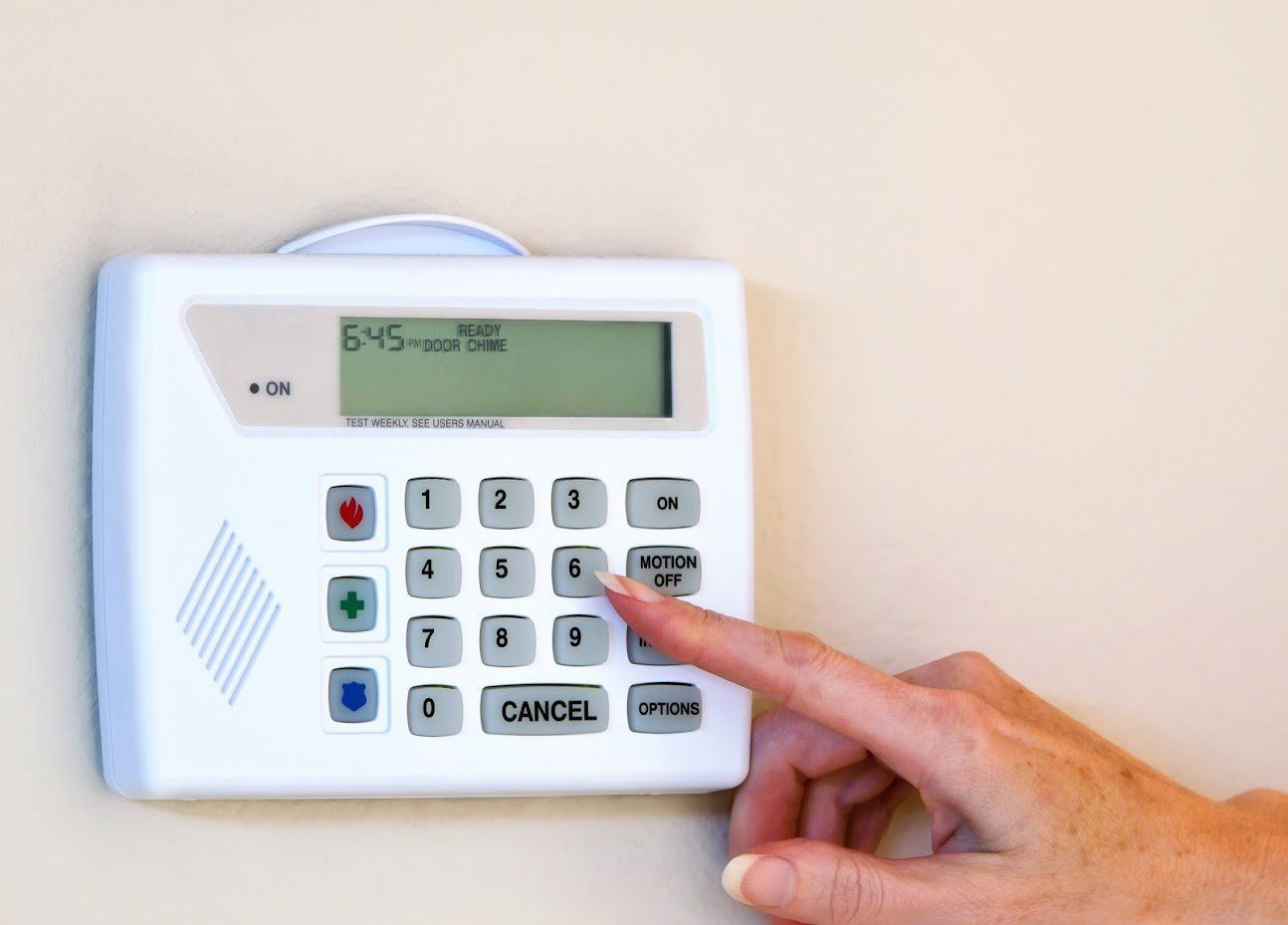 Hand entering code on a white security system keypad mounted on a tan wall. The display reads 
