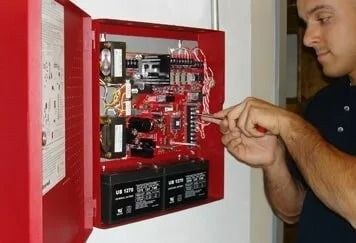 A man is fixing a fire alarm box with a screwdriver. Man working on a red fire alarm control panel mounted on a wall, using a screwdriver. The panel's interior contains wiring, circuit boards, and batteries.