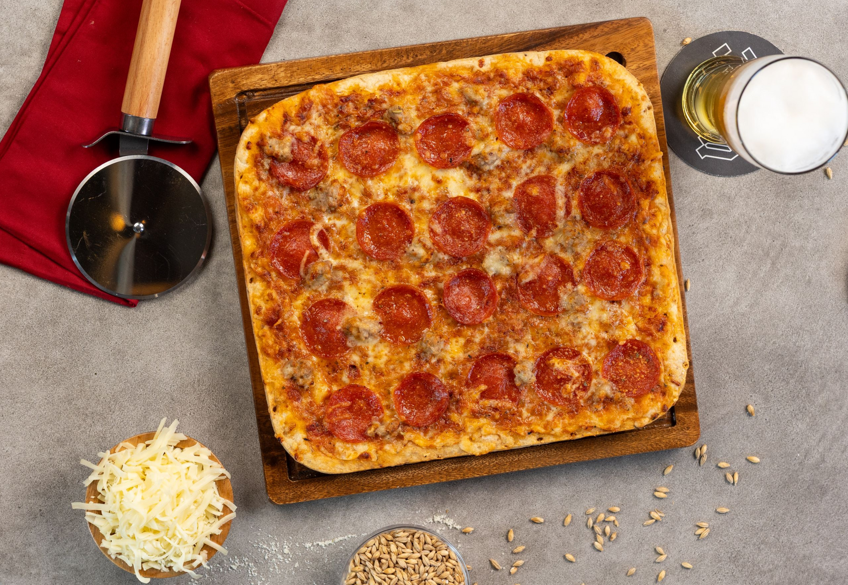 Square pepperoni pizza on a wooden board with cheese, cooking tools, and ingredients on a gray countertop