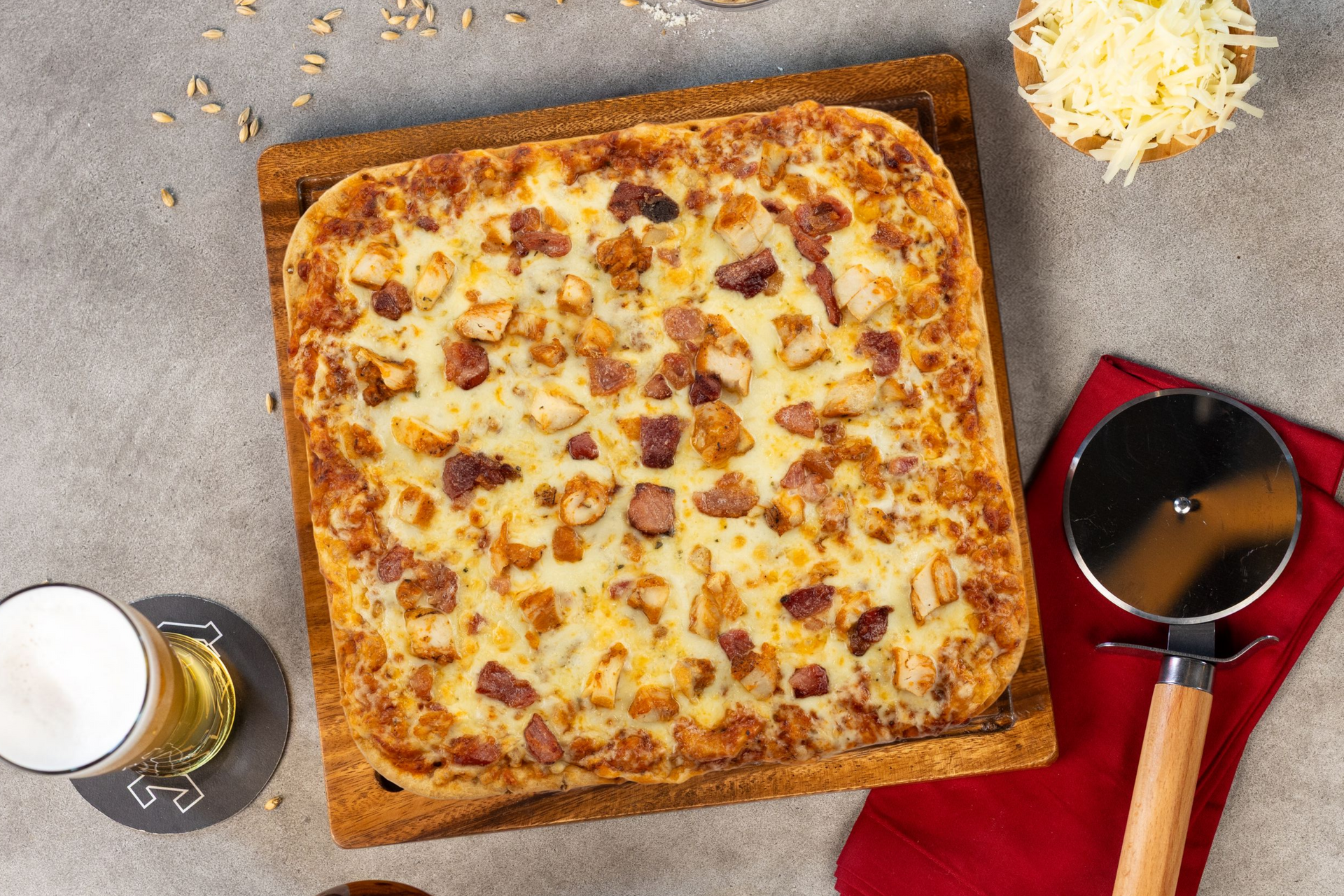 Square cheese pizza on a wooden board with beer, shredded cheese, and a small sauce pot on a red napkin