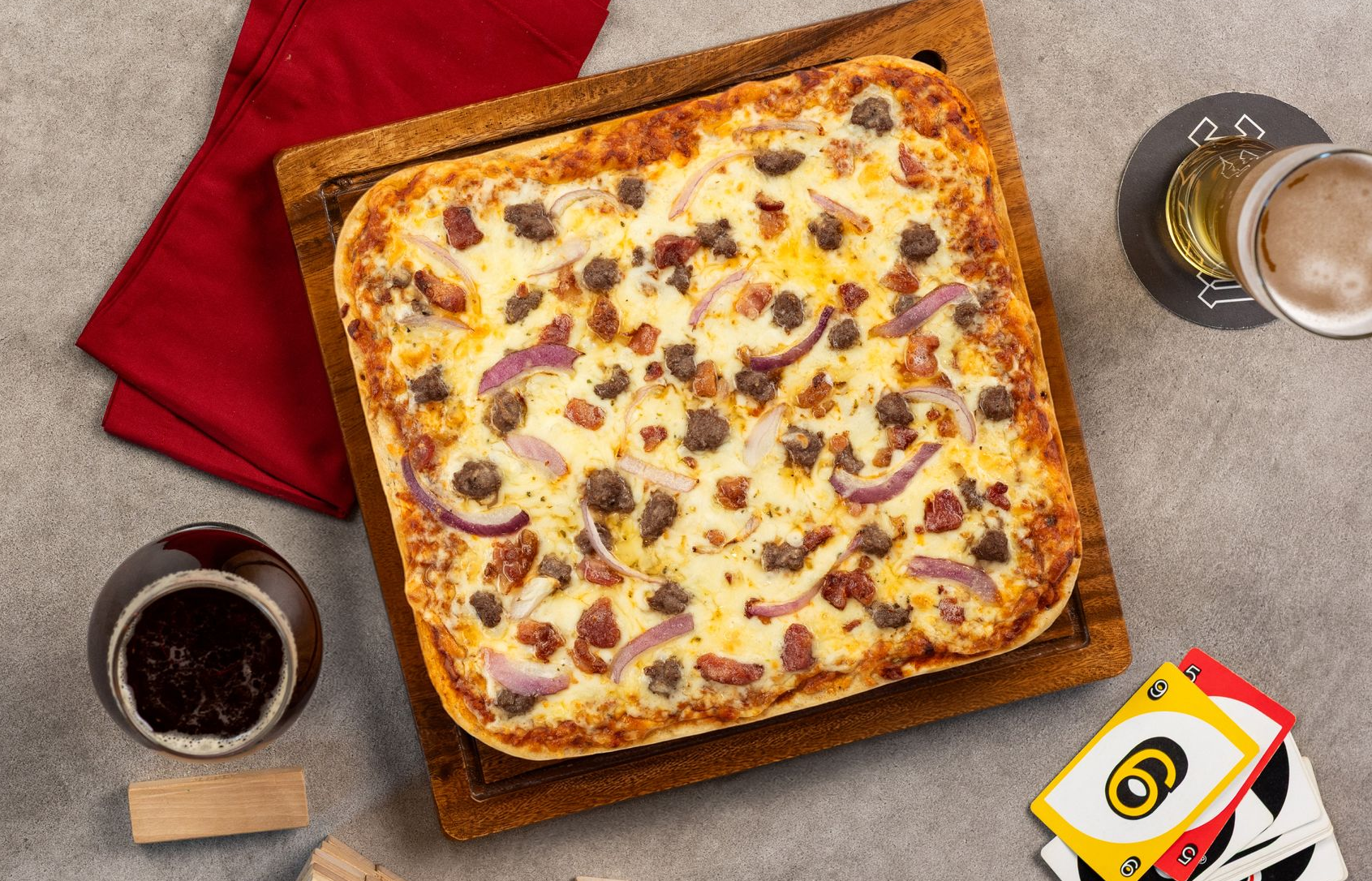 Square pizza on a wooden board with drinks and napkins on a gray table