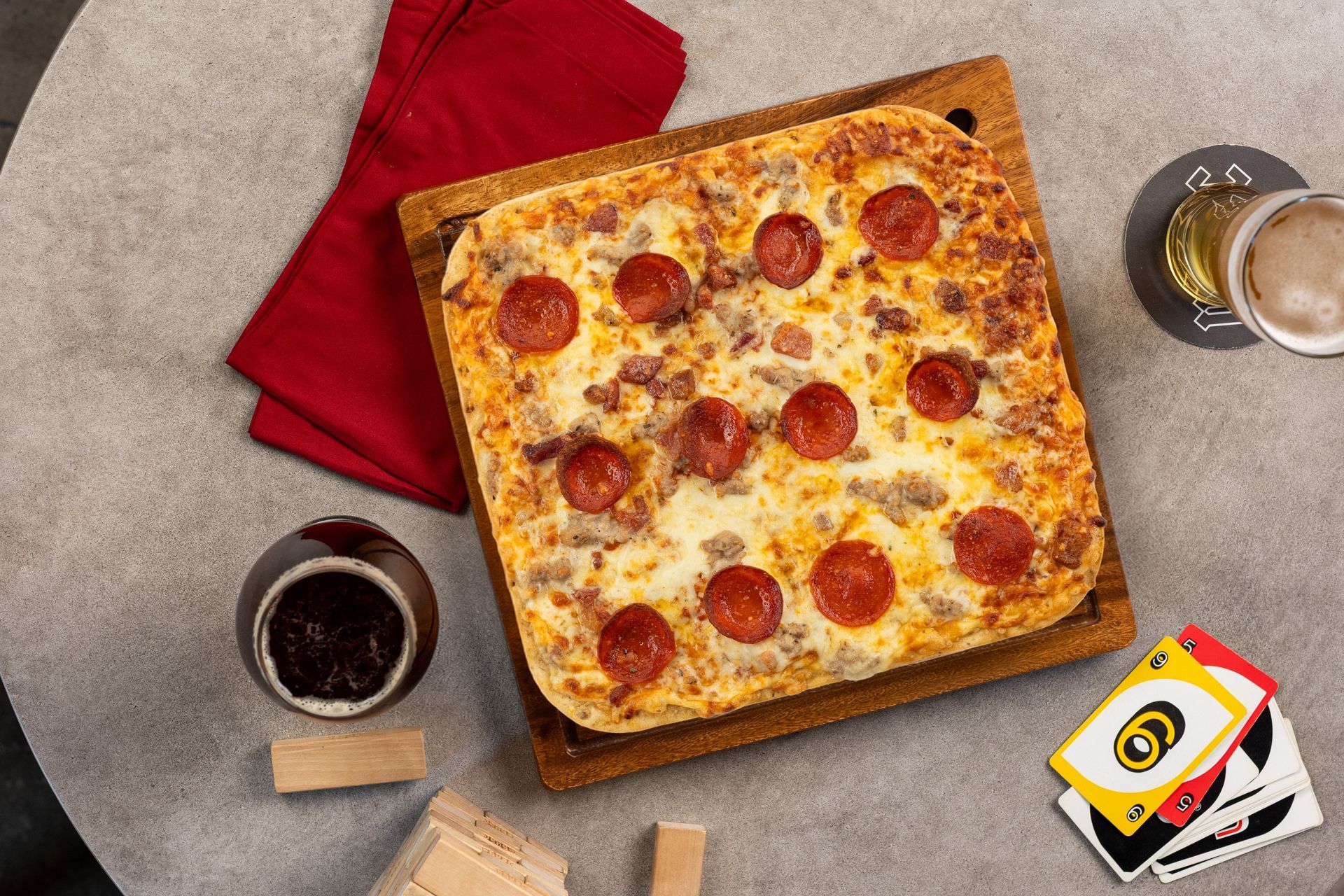 Pepperoni pizza on a wooden board with drinks, napkins, and playing cards on a table