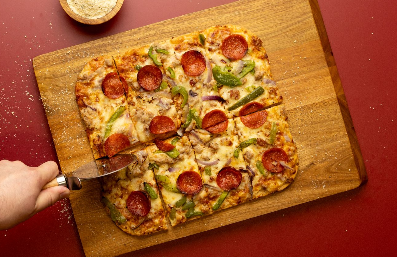 Pepperoni pizza on a wooden board, being cut with a pizza cutter on a red table