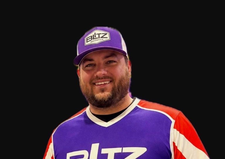 Smiling man in purple and white sports jersey and matching cap on a black background
