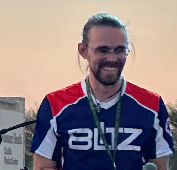 Smiling man in a blue, red, and white jersey with a lanyard, standing at a podium outdoors at sunset