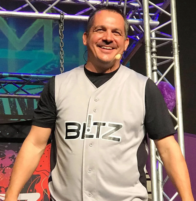 Smiling man in a gray BEIZ vest stands on a colorful stage with metal truss lighting.