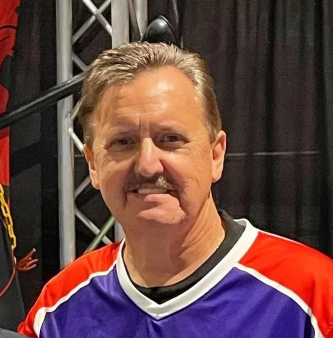 Man wearing a purple sports jersey with orange and white accents, standing in front of a dark backdrop