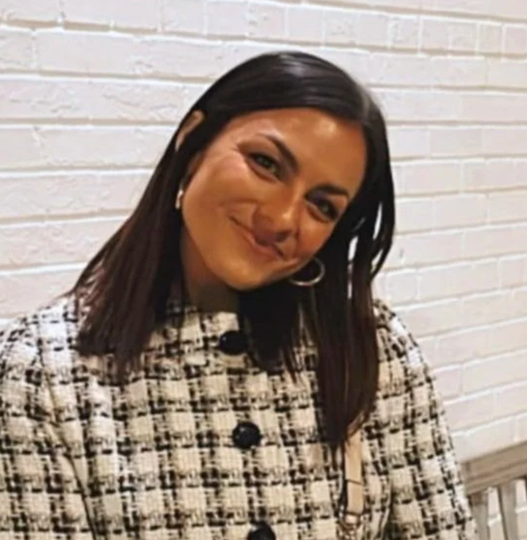 Person smiling in a black-and-white tweed jacket against a white brick wall.