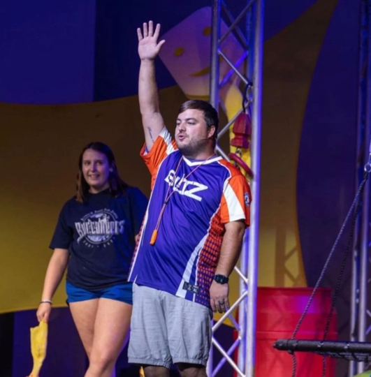 Man on stage in a blue and orange jersey raises one hand; woman stands behind him under purple lighting.