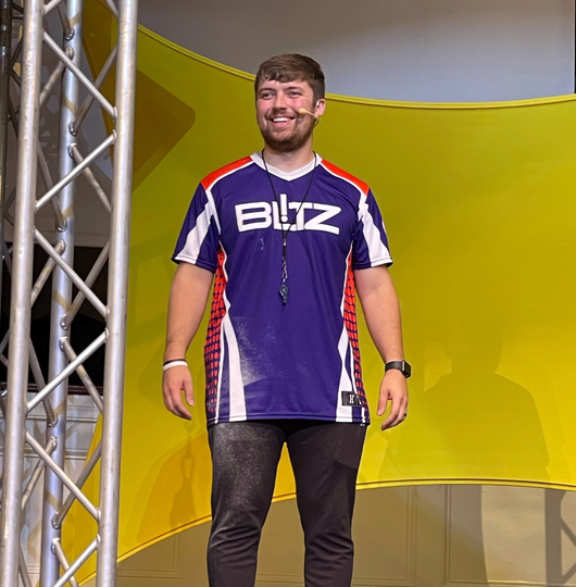 Man wearing a purple BLIZ jersey stands on a stage with a yellow backdrop and metal truss.