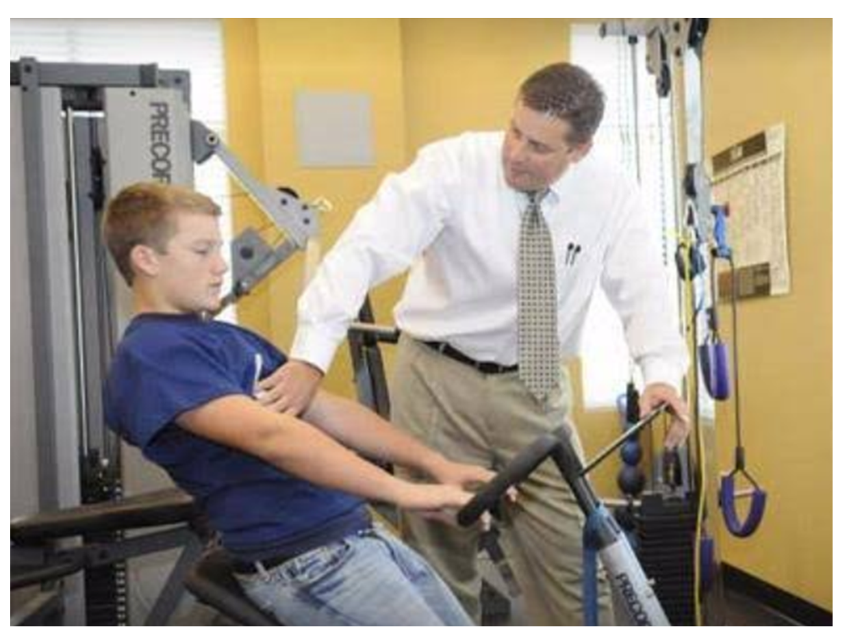 Naperville chiropractor Dr. Whitney guides teen using gym equipment in a yellow-walled room. The teen pulls handles.
