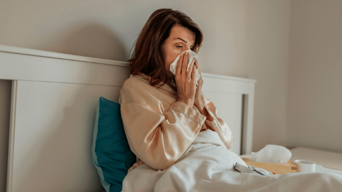 Woman in bed, blowing nose with tissue, appears sick.