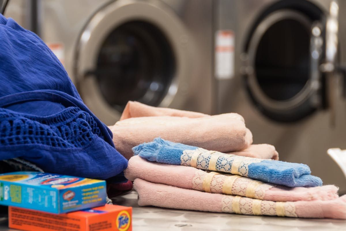 Folded towels and laundry detergent on a table in a laundromat, with washing machines in the background