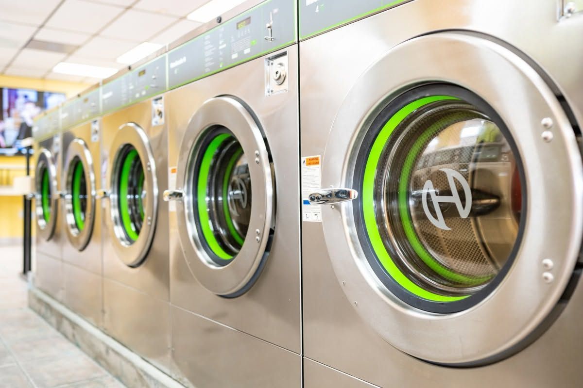Row of stainless steel front-loading washing machines with green-lit doors in a laundromat