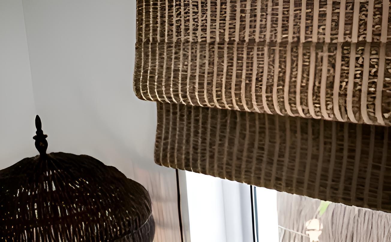 A Wicker Lamp is Sitting Next to a Window With a Roman Shade on It — Lismore Curtains & Blinds In Lismore, NSW