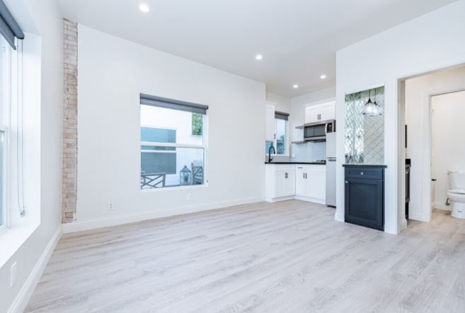 Bright, empty modern living room with white walls, light wood floors, and a small kitchen area.