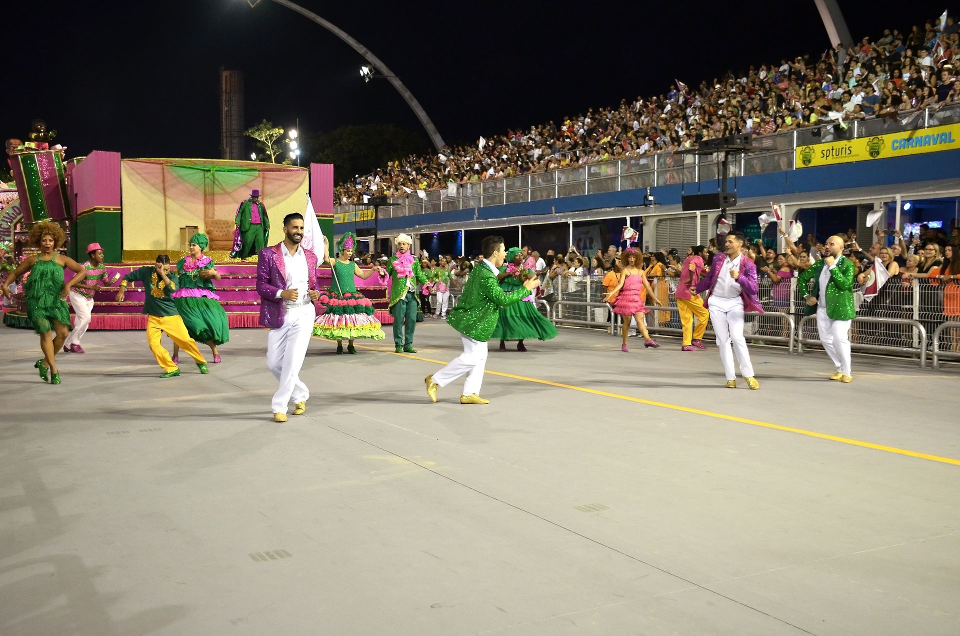 Dançarinos com trajes coloridos se apresentam em um desfile noturno em um estádio com arquibancadas lotadas.