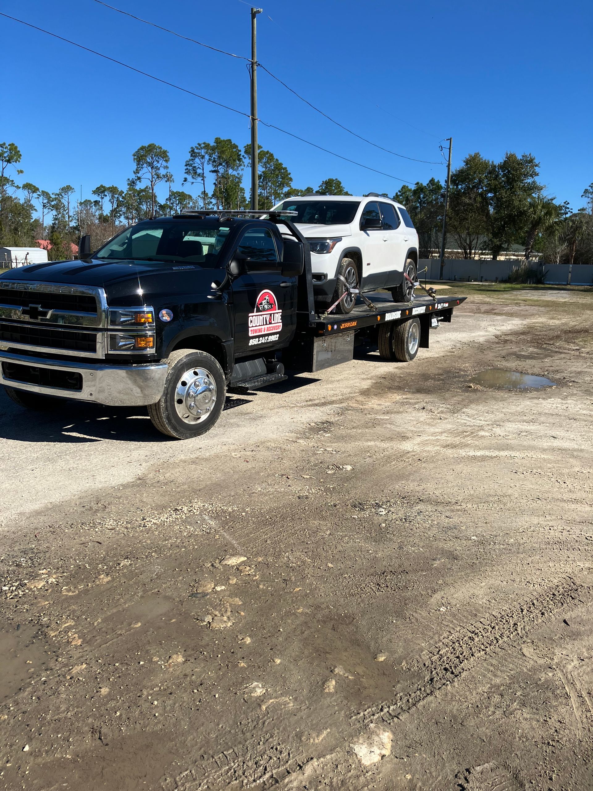 Black tow truck carrying a white SUV on a gravel lot under a blue sky.