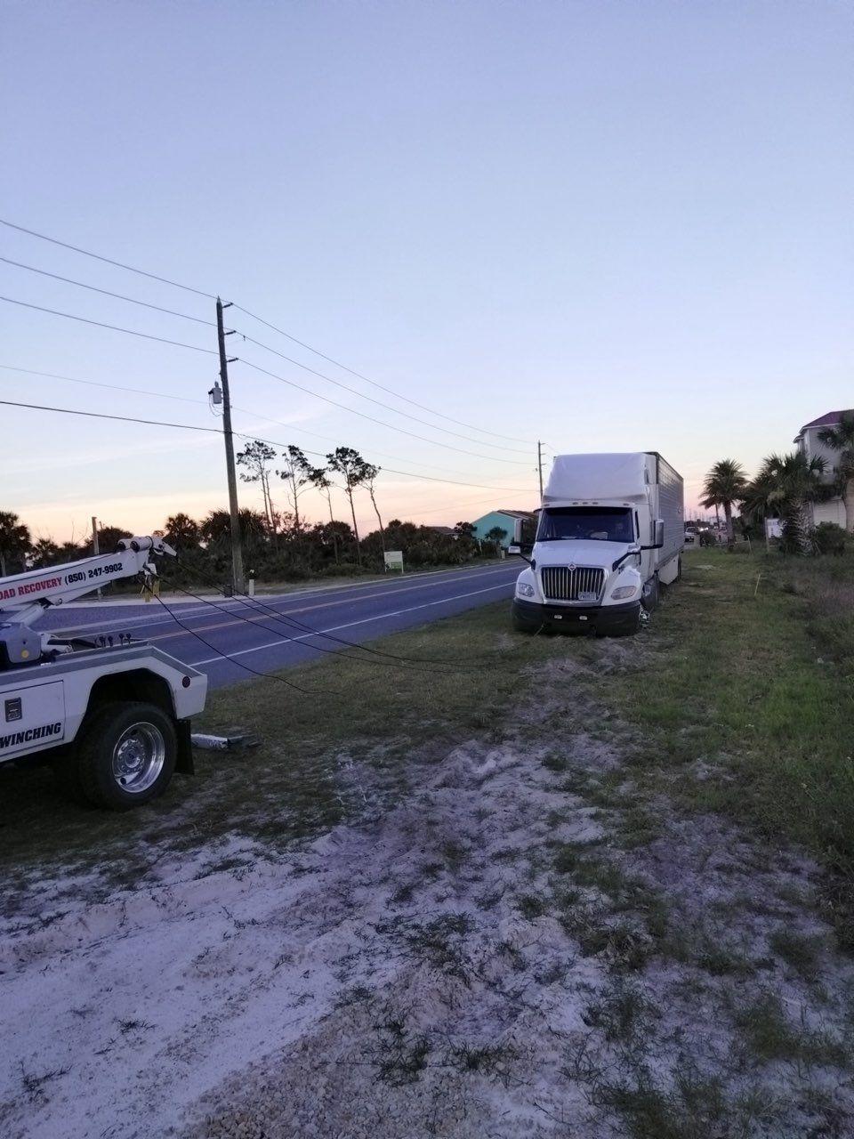 A white semi-truck parked on the grass beside a road, with a tow truck partially visible in the foreground.